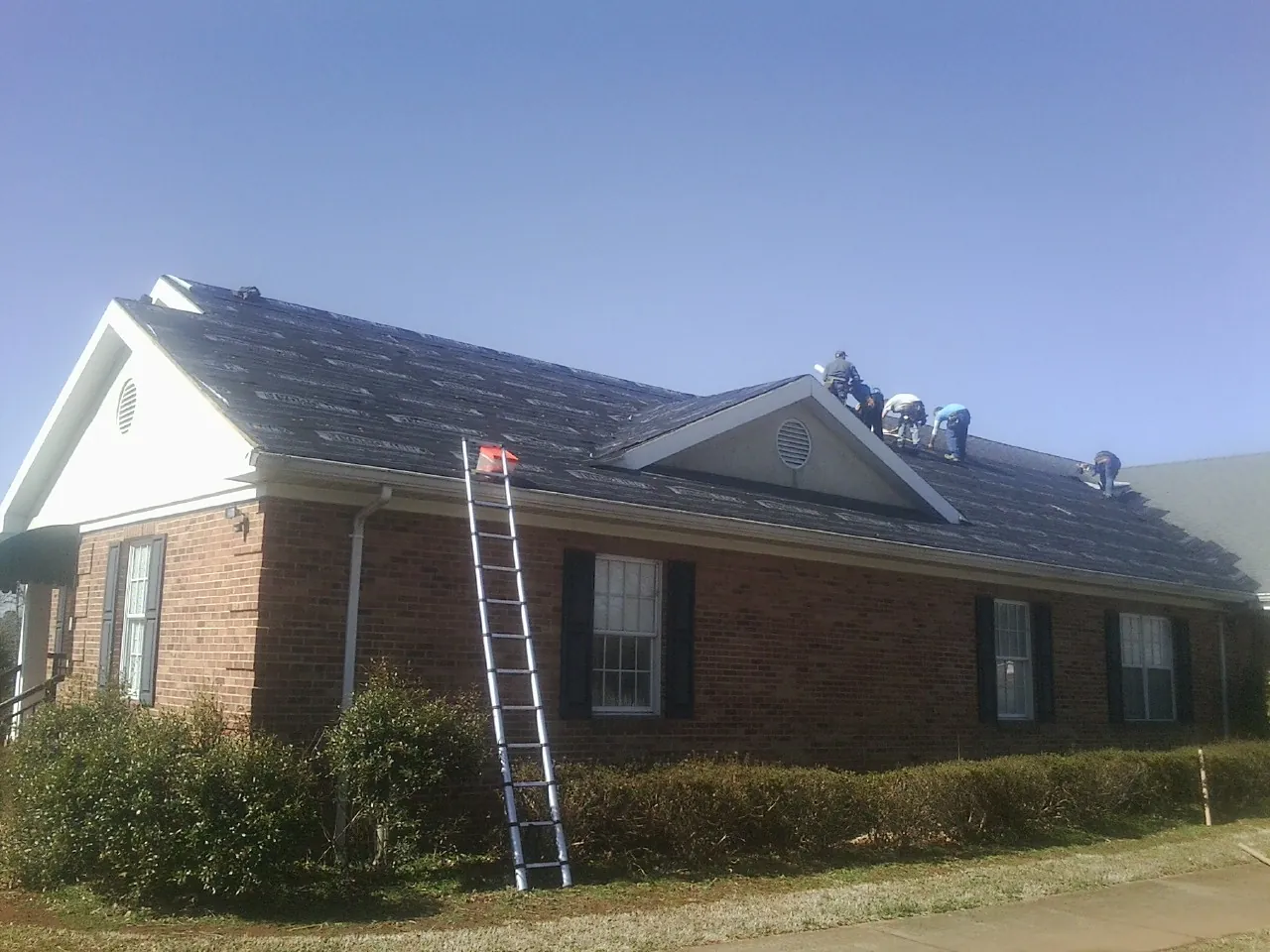 Crew tearing shingles off a brick ranch roof with a ladder set — Emergency Roof Repair work by our crew in Eagle Grove