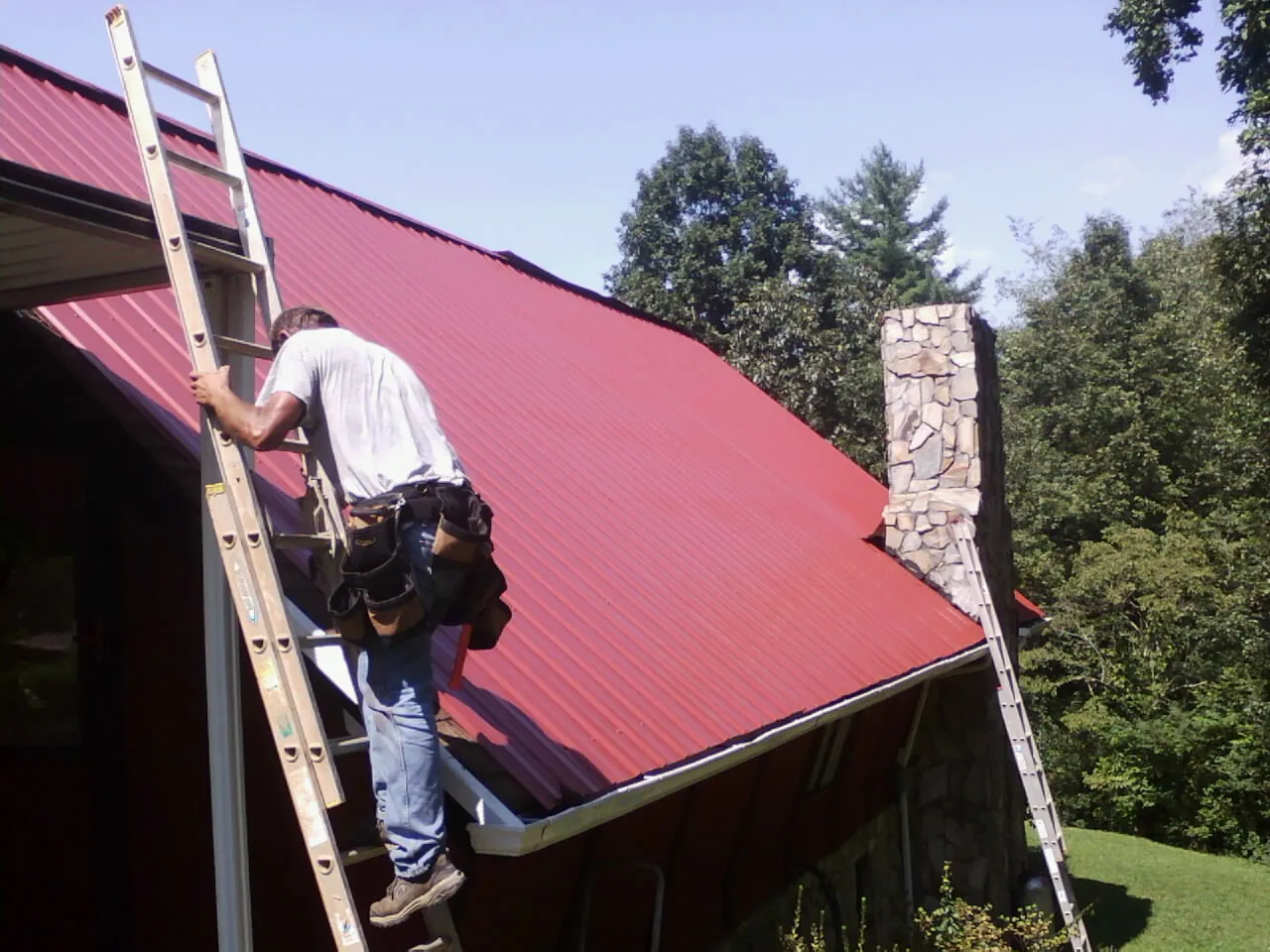 Roofer on a ladder beside a red metal roof and stone chimney — Soffit & Fascia Repair by Roofing Brothers in Eagle Grove